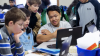 Group of school kids gathered around a laptop, discussing something on the screen.