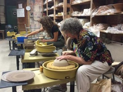 Two femme people sit at ceramics wheels shaping mounds of clay
