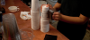 Person stamps a logo onto a stack of white disposable coffee cups on a coffee bar counter.