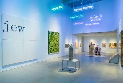 White walled gallery space with 2D work hung on the walls, sculptural work on pedestals in the center of the room, and digital text projection based installations on the upper walls and ceiling.