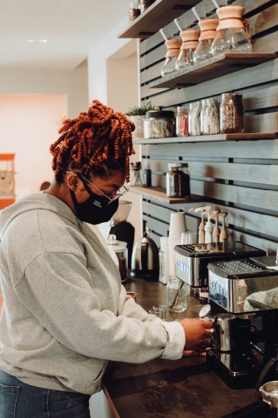 Dark skinned person wearing a surgical face mask makes coffee behind a bar counter