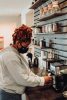 Dark skinned person wearing a surgical face mask makes coffee behind a bar counter