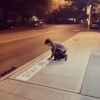 Person kneeling on a sidewalk applies a text based art installation that reads "Stay Dry Out There"