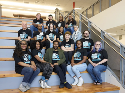 Group of people of different ages, genders, and skin tones sit on bleacher seating, all wearing t-shirts that read "Speak with Purpose"
