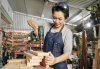 Femme person with short hair wearing a shop apron uses a power drill on a stack of wood boards.