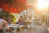 Sunny sidewalk with blue picnic tables and groups of people sitting at them eating and chatting.