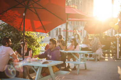 Sunny sidewalk with blue picnic tables and groups of people sitting at them eating and chatting.