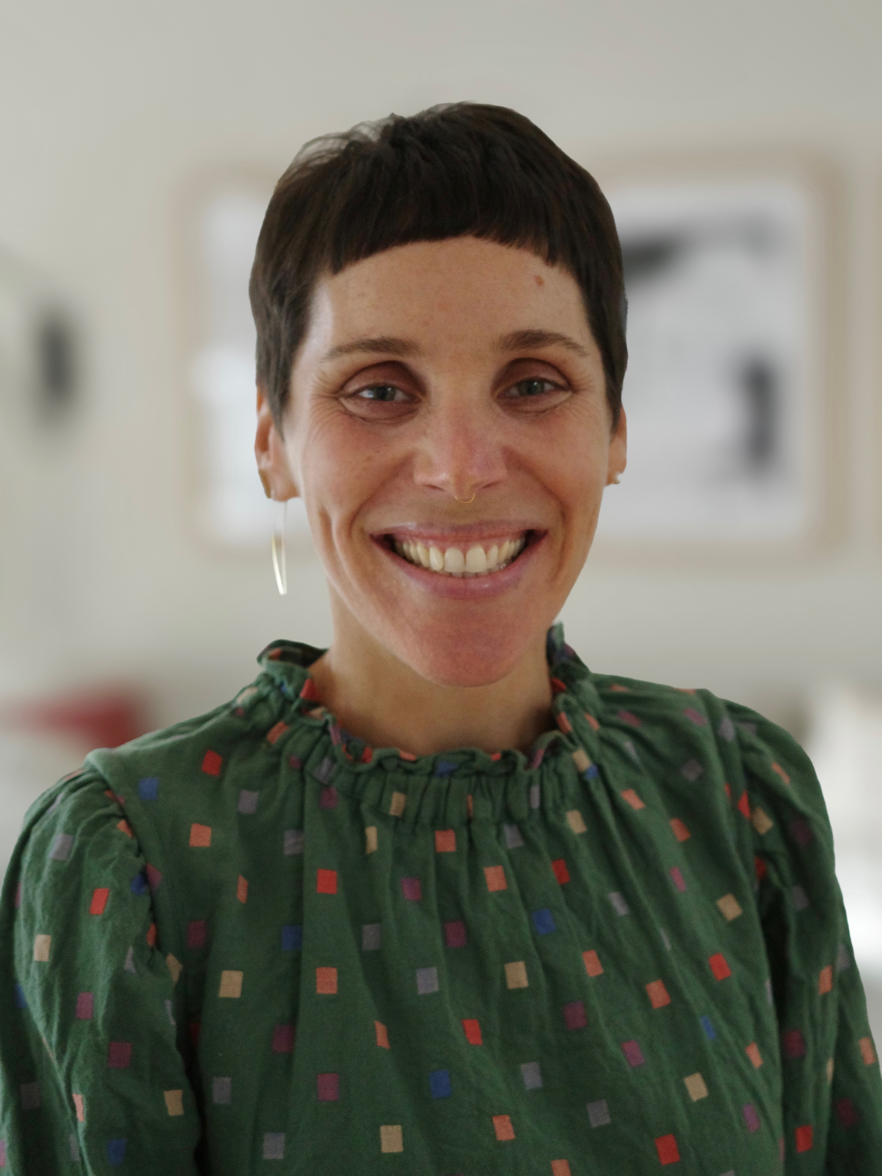 Headshot of Dr. Noa Brazg with short dark hair wearing a green patterned shirt and earrings, photographed indoors against a softly blurred background.