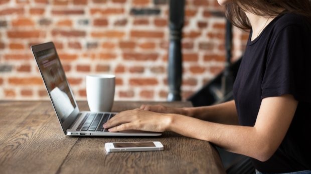 The image shows a person sitting at a wooden table, using a laptop. A white coffee mug is placed next to the laptop, and a smartphone is lying on the table in front. The background consists of a brick wall with a black railing or pillar partially visible.