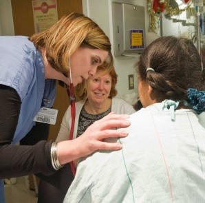 A healthcare worker in scrubs is leaning forward and placing a supportive hand on a pediatric patient's shoulder. The scene takes place in a medical setting, another person visible in the background.