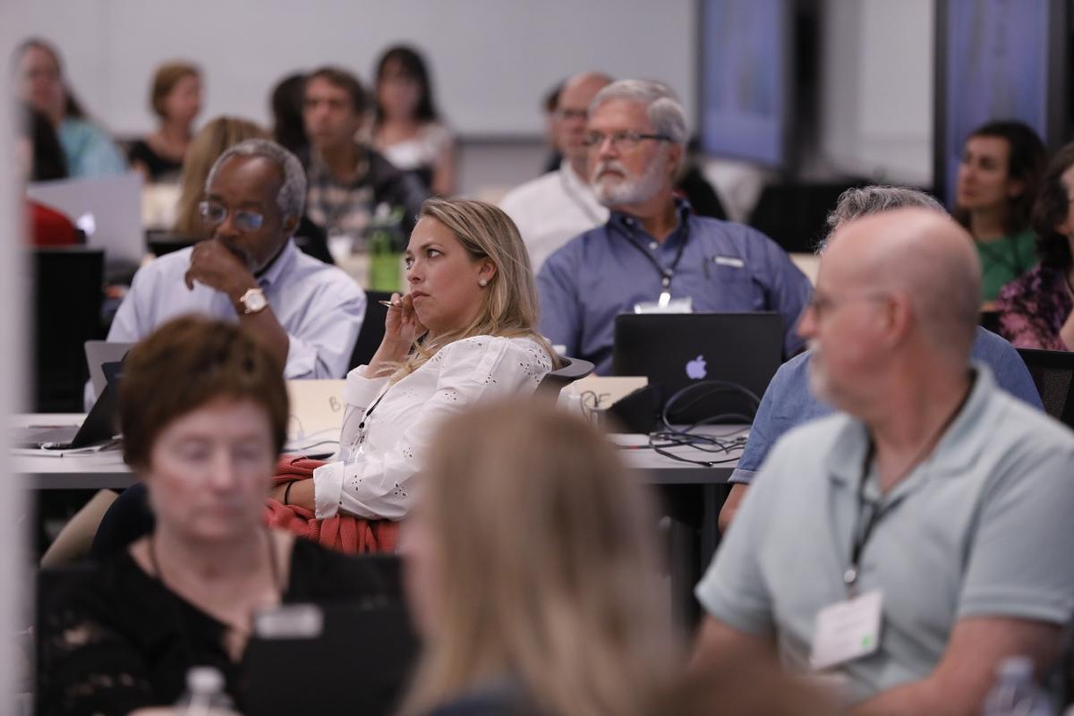 A group of people are seated in a conference or classroom setting, attentively listening to a presentation. Laptops, notebooks, and other materials are placed on the tables. The background shows multiple display screens and a professional environment.