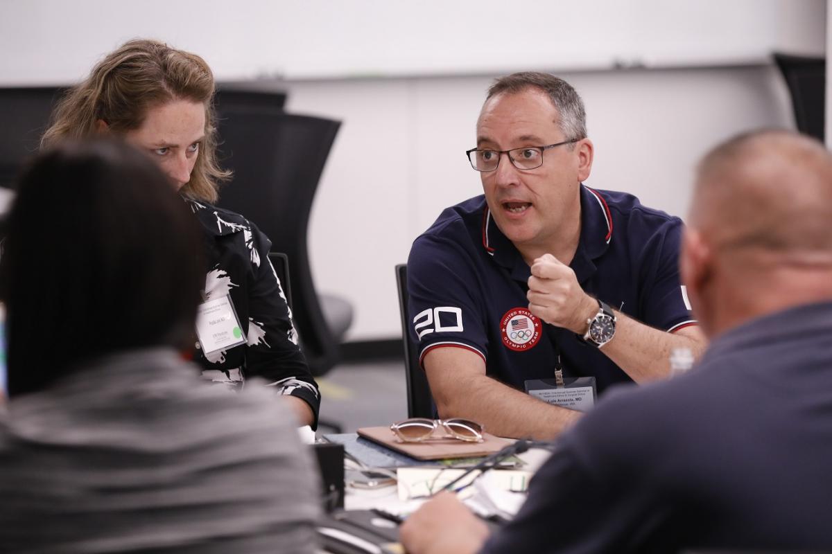 A group of people are seated around a table engaged in discussion. One person is wearing a navy blue shirt. Items such as a pair of glasses and notebooks are on the table. The setting appears to be a meeting or workshop in a conference room.