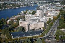 Aerial view of a UW Medical Center -Montake - large urban campus beside lake washington, with multiple mid-rise buildings, tree-lined streets, and a major bridge spanning the water in the background. Cars travel along a broad road on the right, and marinas and neighborhoods line the riverbanks.