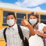 Three students wearing masks stand in front of a school bus. One gives a thumbs up.
