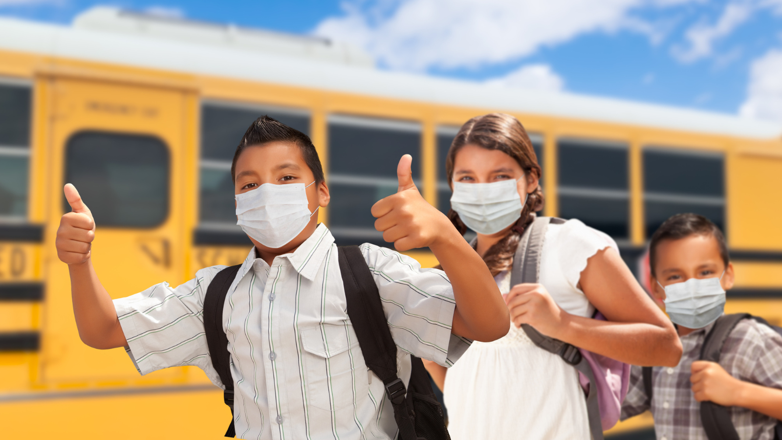 Three students wearing masks stand in front of a school bus. One gives a thumbs up.