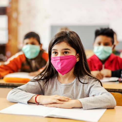 A young girl sits in class attentively while wearing a mask.