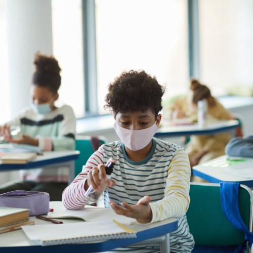 Student sits in class with mask on and sprays hand sanitizer into his hand.