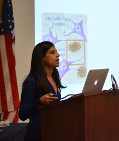 A woman with dark hair speaks at a podium in front of a slide showing brain cells