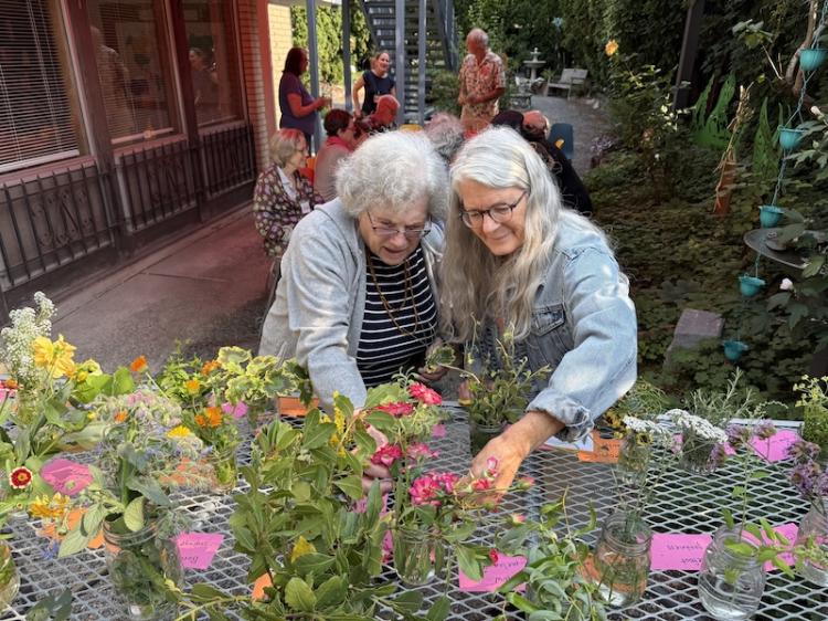 Two women reach to flowers on a table, with a view of a people in a garden behind them