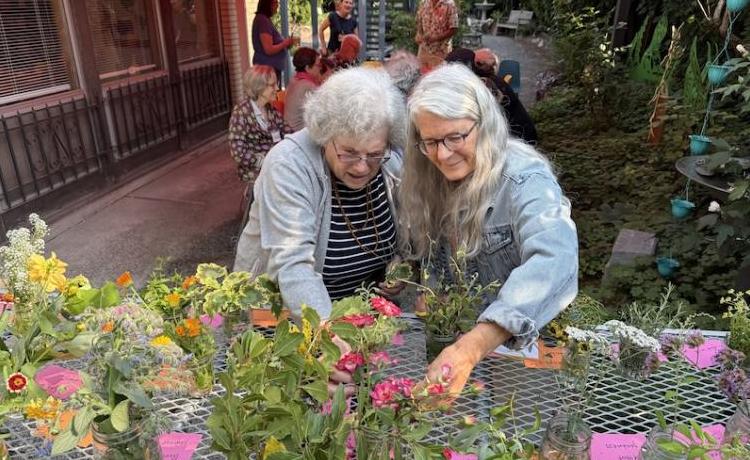 Two women reach to flowers on a table, with a view of a people in a garden behind them