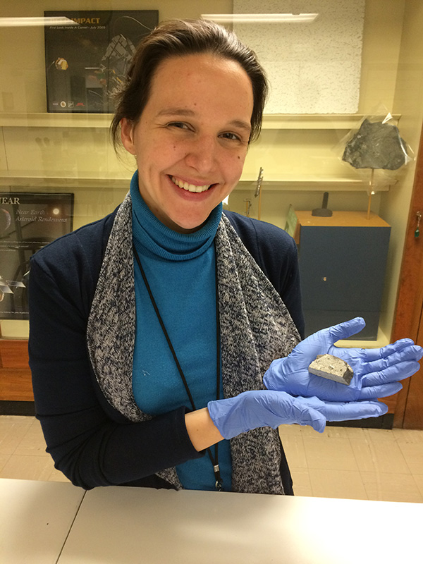 Dr. Nicole Lunning holds a piece of meteorite. She is a white women with brown hair (pulled back) wearing a blue turtleneck under a blue cardigan sweater. A small chunk of grey-colored meteorite rests in her blue-glove-covered hands.