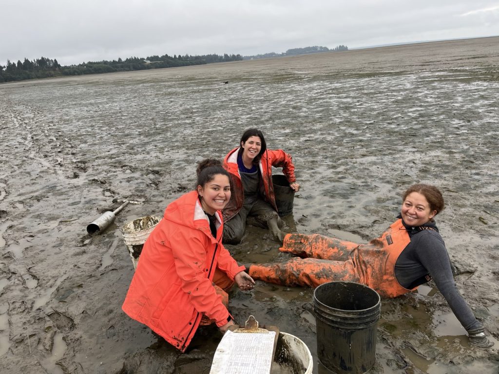 Three scientists wearing water-proof bright orange clothes are covered in mud, sitting on a mud flat. Equipment is scattered around them. They smile at the camera. 