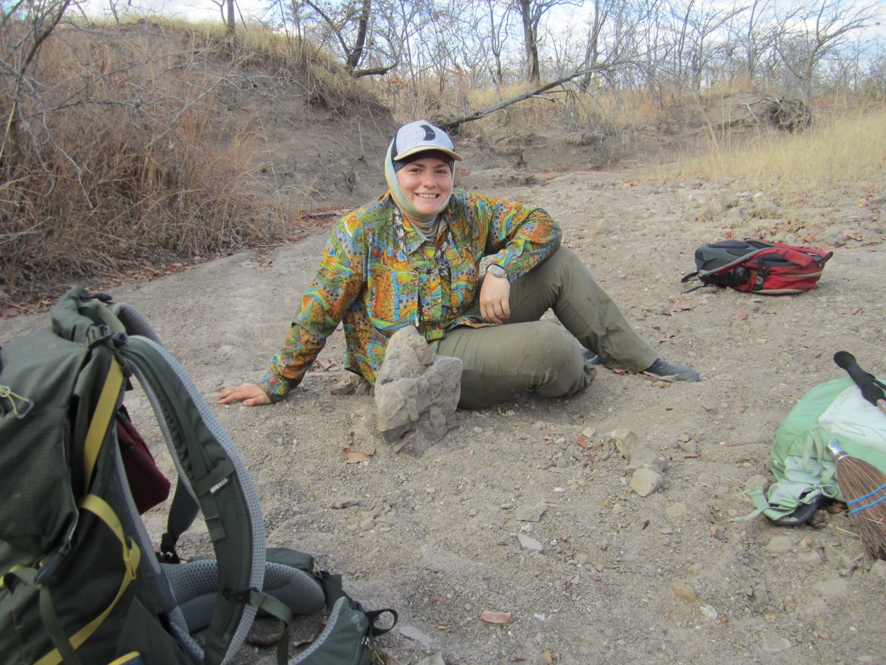 A scientist in field clothes sits behind a basket ball sized fossil. 