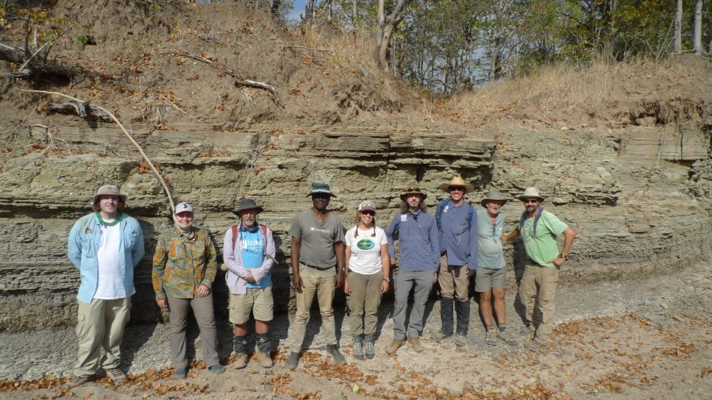 Nine people in field clothes stand in front of a rock outcrop and smile at the camera. 