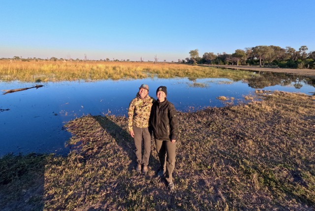 Two people smile at the camera in front of a flooded grassland. 