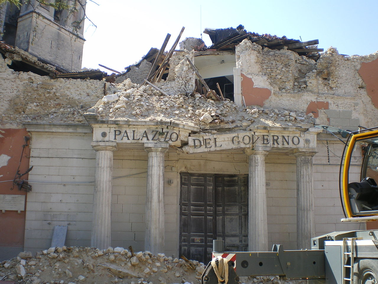 A collapsed building with the words "Palazzo Del Governo" on a broken edifice supported by four pillars.