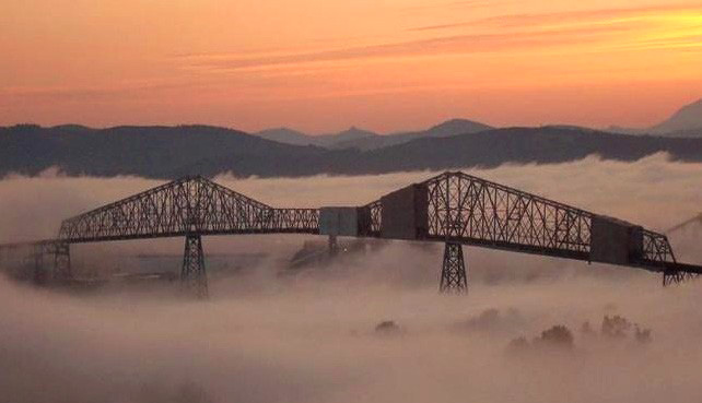 Photo of a bridge with clouds or fat below and mountains in the distance at sunset.