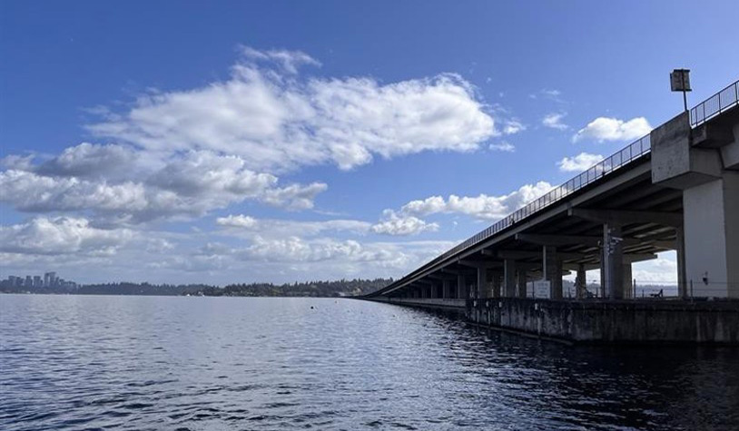 View of Homer Hadley Floating Bridge across Lake Washington, between Mercer Island and Seattle, Washington state, from a lake-level perspective. Part of the concrete bridge support structure is visible under the bridge deck. In the distance, cityscape and trees are seen against a background of blue sky and clouds.