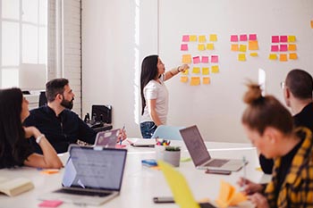 Business meeting with 4 people sitting at a table listening to a woman at the board talking while pointing at post-it notes.