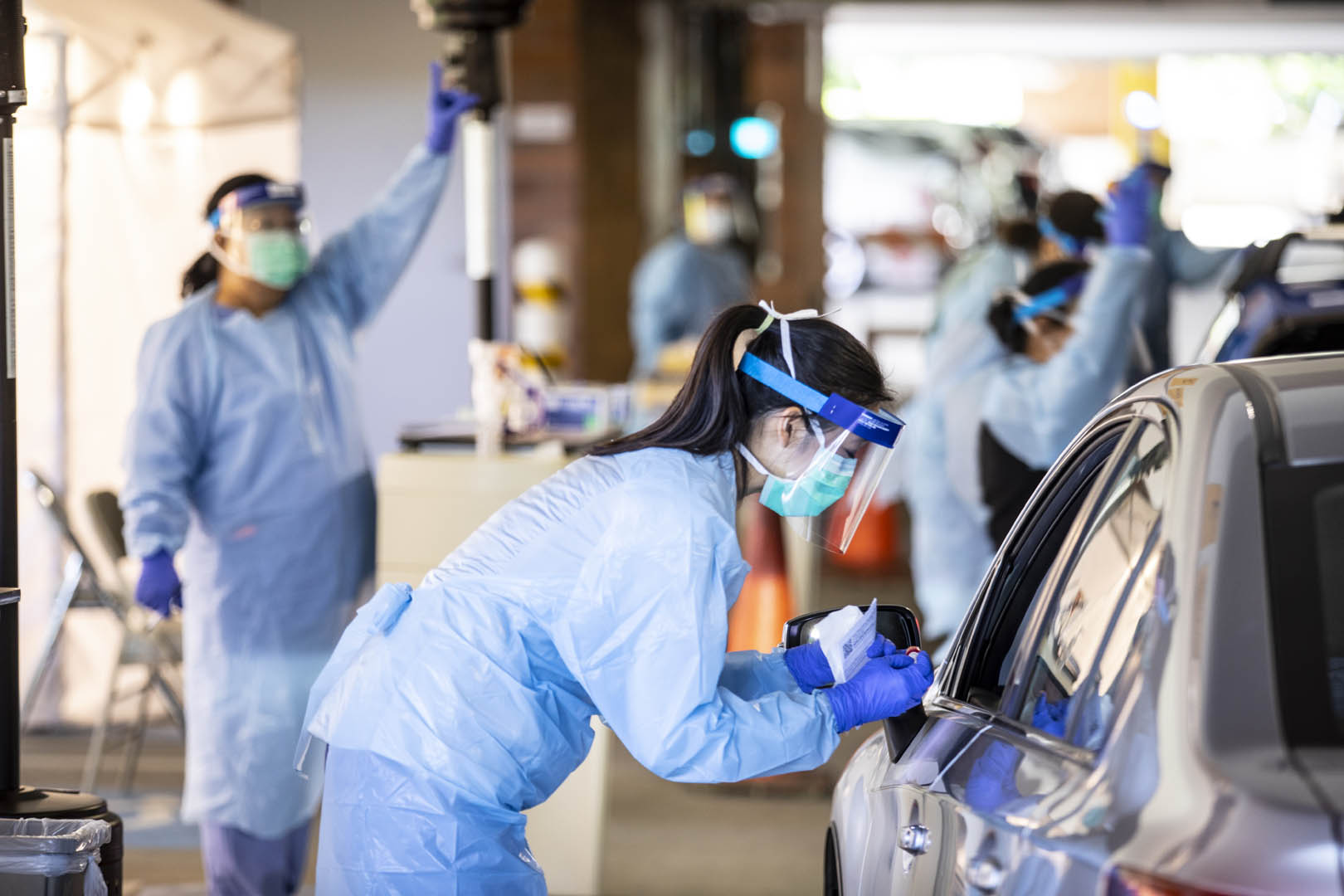 Nurses administering COVID tests at a drive-up testing facility. A nurse in the foreground is talking to a driver and a nurse in the background is raising her hand to let the next vehicle know they should move forward.