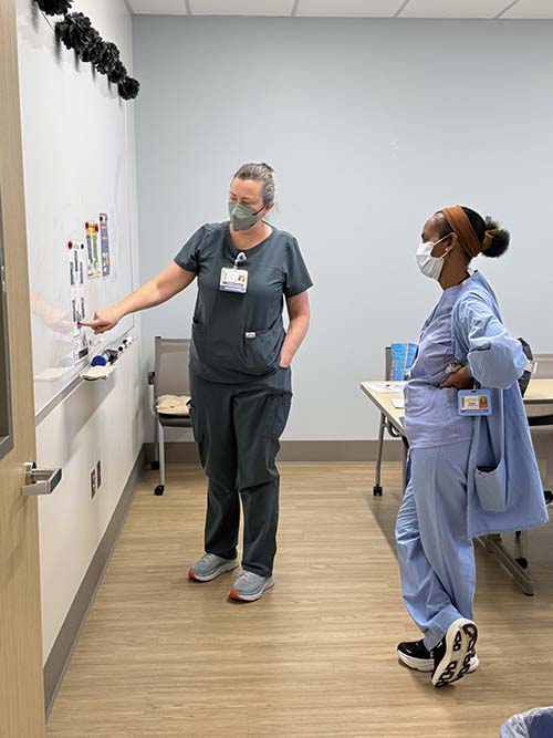 Two masked nurses looking at a whiteboard together. One nurse is pointing at the whiteboard.