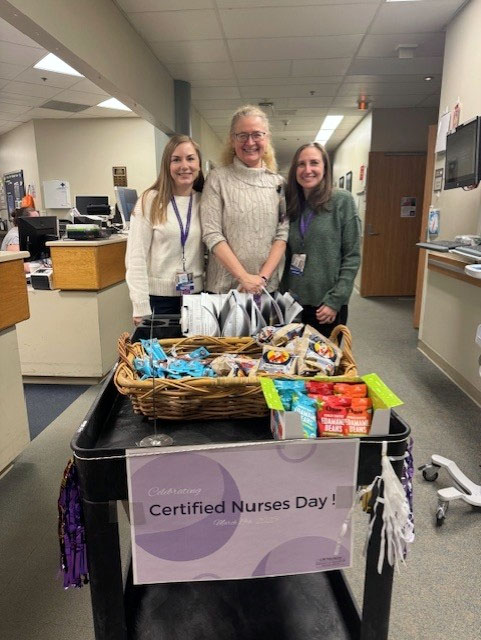 Three nurses posing near a unit nursing station near a cart full of treats with a sign that says Celebrating Certified Nurses Day.